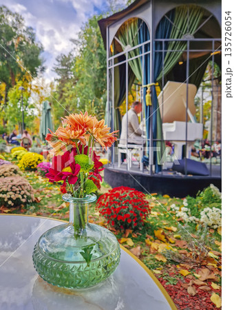 Charming outdoors cafe scene with focus to a vibrant vase of flowers on the table and a gazebo with a white piano in the background. A pianist performing live music in the colorful autumn square 135726054
