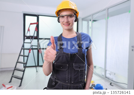 Confident woman in safety gear gives a thumbs up while working on a construction site in a bright, modern building 135726141