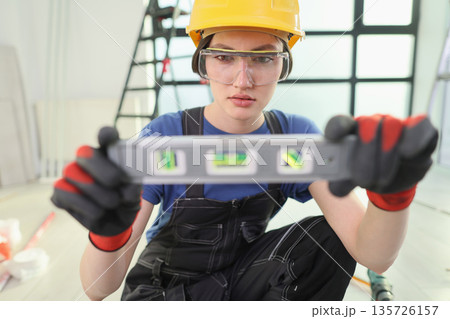Young construction worker carefully checks wall level in bright workshop while wearing safety gear and focused expression Young construction worker carefully checks wall level in bright workshop while wearing safety gear and focused expression 135726157