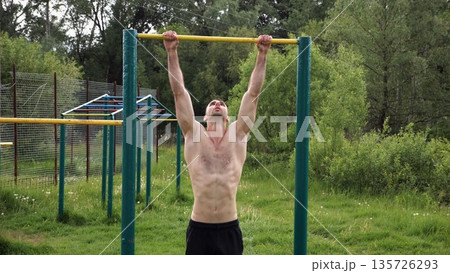 Muscular athlete doing pull ups on horizontal bar at sports ground. Young shirtless sportsman exercising at nature. Strong sporty man training outdoor. Concept of healthy and active lifestyle. Slow mo Muscular athlete doing pull ups on horizontal bar at sports ground. Young shirtless sportsman exercising at nature. Strong sporty man training outdoor. Concept of healthy and active lifestyle. Slow mo 135726293