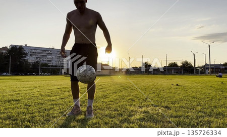 Young man juggling soccer ball on stadium at sunset. Professional footballer kicking ball at green field. Sportsman practicing tricks at meadow with sunlight at background. Freestyle football 135726334