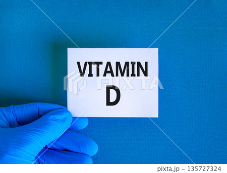 Vitamin D symbol. Concept words Vitamin D on beautiful white card. Doctor's hand in blue medical glove. Beautiful blue background. Medical healthy lifestyle Vitamin D concept. Vitamin D symbol. Concept words Vitamin D on beautiful white card. Doctor's hand in blue medical glove. Beautiful blue background. Medical healthy lifestyle Vitamin D concept. 135727324