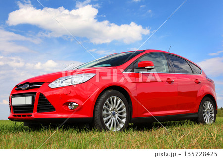 Red car on a blue sky background with white clouds. The car is standing in the field on the green grass. Red car on a blue sky background with white clouds. The car is standing in the field on the green grass. 135728425