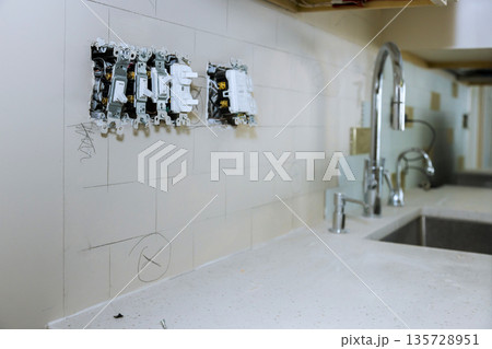 Workers are installing electrical outlets on wall of kitchen, wall has white tiles, sink faucet are visible nearby. Workers are installing electrical outlets on wall of kitchen, wall has white tiles, sink faucet are visible nearby. 135728951
