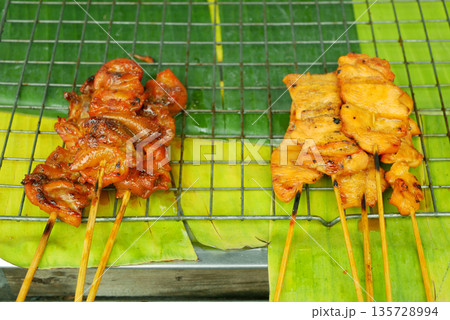 Meat skewer street food vendor displaying assorted grilled like raw chicken skin close-up detail, heart, gizzard, liver and pork meat skewers on banana at busy street meal stall. Food barbecue or Thai Meat skewer street food vendor displaying assorted grilled like raw chicken skin close-up detail, heart, gizzard, liver and pork meat skewers on banana at busy street meal stall. Food barbecue or Thai 135728994