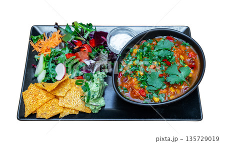 Colorful vegetarian chili served in a bowl with fresh salad, tortilla chips, and guacamole on a black plate, showcasing a vibrant meal presentation Colorful vegetarian chili served in a bowl with fresh salad, tortilla chips, and guacamole on a black plate, showcasing a vibrant meal presentation 135729019