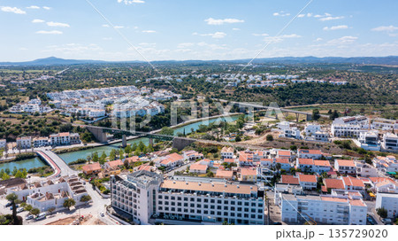 This aerial view showcases Tavira, a town in the Algarve region, with buildings, a river, and the surrounding landscape visible under clear skies This aerial view showcases Tavira, a town in the Algarve region, with buildings, a river, and the surrounding landscape visible under clear skies 135729020