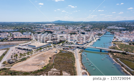 Tavira in Algarve Portugal with buildings along the Gilao river and green areas. This is a sunny day with boats in the water and hills in the background Tavira in Algarve Portugal with buildings along the Gilao river and green areas. This is a sunny day with boats in the water and hills in the background 135729094
