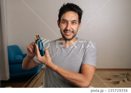 Portrait of handsome bearded young man holding teal and gold bottle, presenting skincare product smiling looking to camera. Concept of male grooming, beauty, advertising. Portrait of handsome bearded young man holding teal and gold bottle, presenting skincare product smiling looking to camera. Concept of male grooming, beauty, advertising. 135729356