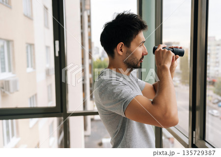 Side view of voyeurist male curiously peering out window using monocular, watching with intense emotion. Engaging in voyeurism. Observing surveillance. Peeping through curtains. Maintaining vigilance. Side view of voyeurist male curiously peering out window using monocular, watching with intense emotion. Engaging in voyeurism. Observing surveillance. Peeping through curtains. Maintaining vigilance. 135729387