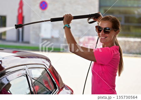 Young woman wearing pink t shirt and sunglasses cleaning her car in self service carwash, spraying water over front windshield and roof Young woman wearing pink t shirt and sunglasses cleaning her car in self service carwash, spraying water over front windshield and roof 135730564