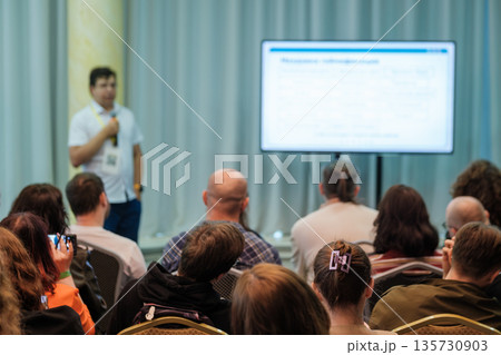 Presenter speaks at conference to attentive audience with large screen and notes during professional seminar Presenter speaks at conference to attentive audience with large screen and notes during professional seminar 135730903