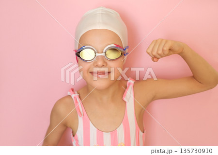 A cheerful girl in a swimming cap and mirrored glasses on a pink studio background shows off her strong and sturdy biceps 135730910