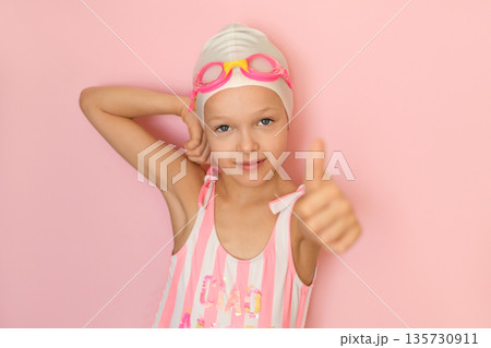 A girl in a swimming cap and glasses on a pink studio background shows a thumbs-up gesture. 135730911
