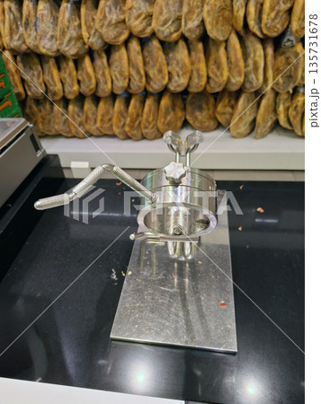 A metal device holds a leg of Spanish jamon ham securely in place at a supermarket. Staff use it to slice the ham for customers. The setup is in a food section with other products nearby. A metal device holds a leg of Spanish jamon ham securely in place at a supermarket. Staff use it to slice the ham for customers. The setup is in a food section with other products nearby. 135731678