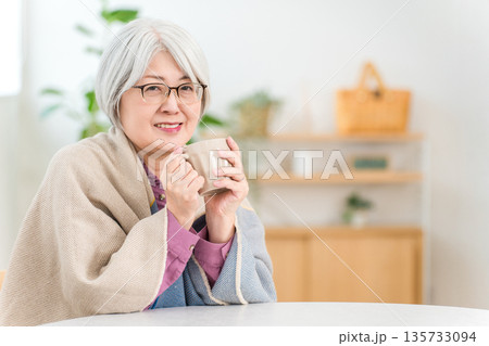 リビングで肩から毛布をかけ寒がるシニア女性（ホットドリンク,温まる,温活） 135733094
