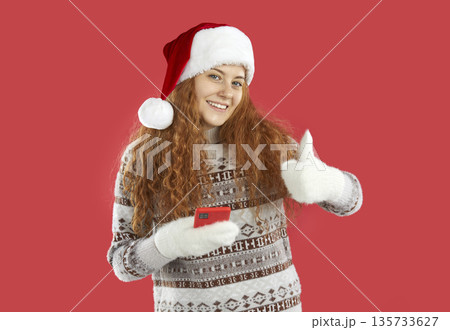 Pretty redhead Christmas girl in Santa hat showing thumb up holding smartphone on red background. Pretty redhead Christmas girl in Santa hat showing thumb up holding smartphone on red background. 135733627