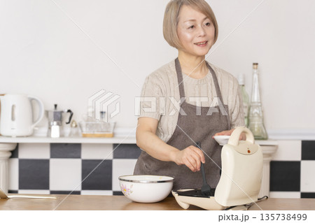 キッチンでアメリカンワッフルを焼くシニア女性 キッチンでアメリカンワッフルを焼くシニア女性 135738499