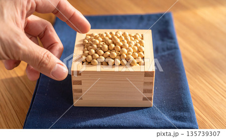 陽光が差し込む木目のテーブルで大豆が入った枡に手を伸ばす節分の風景 135739307