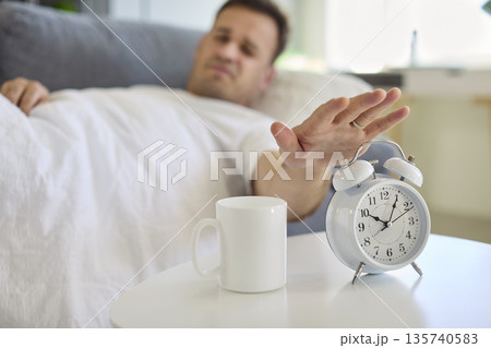 Sleepy man reaching to alarm clock in bed, morning wake-up with coffee mug 135740583