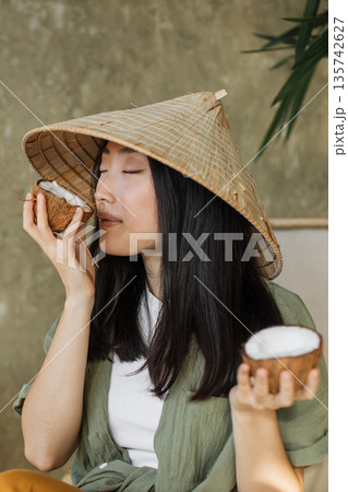 A young Asian woman wearing a traditional conical hat enjoys the scent of fresh coconut halves A young Asian woman wearing a traditional conical hat enjoys the scent of fresh coconut halves 135742627