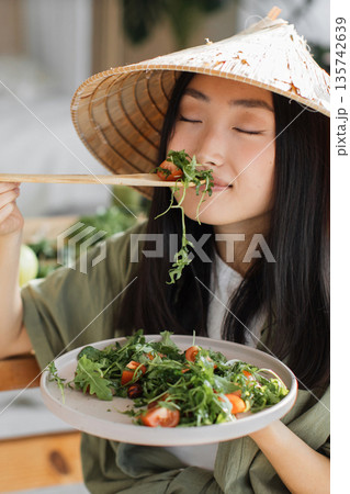 A young Asian woman wearing a conical hat enjoys a fresh salad with cherry tomatoes and greens, bringing a bite to her mouth with chopsticks A young Asian woman wearing a conical hat enjoys a fresh salad with cherry tomatoes and greens, bringing a bite to her mouth with chopsticks 135742639
