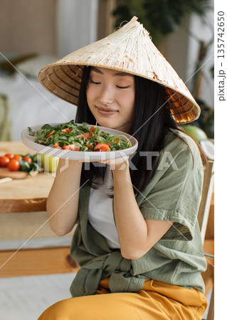 A young woman wearing a conical hat savors the aroma of a freshly prepared salad with tomatoes and greens 135742650