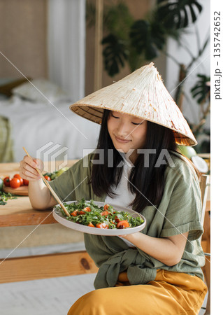 A young Asian woman wearing a conical hat enjoys a fresh salad with chopsticks at a wooden table, surrounded by fresh ingredients A young Asian woman wearing a conical hat enjoys a fresh salad with chopsticks at a wooden table, surrounded by fresh ingredients 135742652
