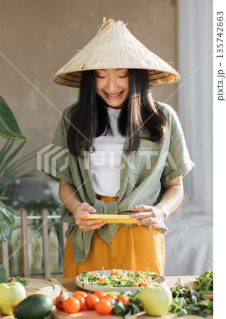 A young Asian woman wearing a conical hat smiles while preparing a fresh salad with various ingredients on a wooden table A young Asian woman wearing a conical hat smiles while preparing a fresh salad with various ingredients on a wooden table 135742663