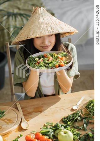 A young woman wearing a conical hat savors the aroma of a freshly prepared salad filled with greens and tomatoes 135742668