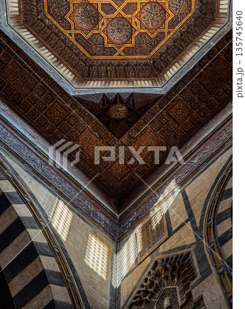 Interior view of Sultan Al-Ashraf Qaytbay Mosque and mausoleum. Features intricate arches, striped wall, and decorative dome. Soft light filters through stained glass window 135745640