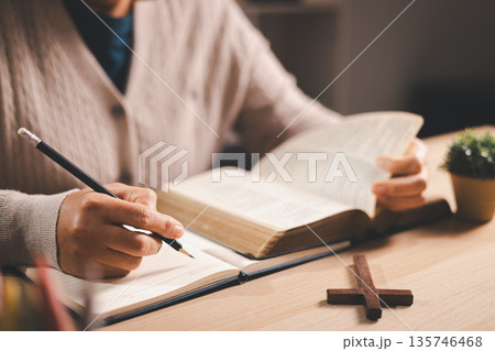 Person taking notes while reading an open Bible at a desk, pencil and journal beside a wooden cross, warm indoor light and soft focus, conveying Christian faith, devotion and scripture study at home. Person taking notes while reading an open Bible at a desk, pencil and journal beside a wooden cross, warm indoor light and soft focus, conveying Christian faith, devotion and scripture study at home. 135746468