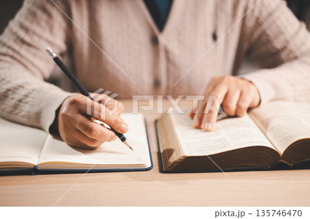 Hands taking notes in a notebook while studying an open Bible, finger tracking the text, warm indoor light and soft focus, conveying faith, devotion, learning and reflection at home. 135746470