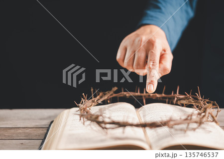 Hand pointing to a crown of thorns on an open Bible over rustic wood, dramatic light and dark background with copy space, symbolizing crucifixion, sacrifice, Holy Week, Good Friday and Easter faith. Hand pointing to a crown of thorns on an open Bible over rustic wood, dramatic light and dark background with copy space, symbolizing crucifixion, sacrifice, Holy Week, Good Friday and Easter faith. 135746537
