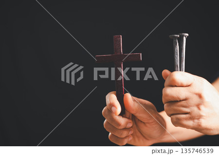 Hands holding a wooden cross and two iron nails on dark background, dramatic light and copy space, symbolizing crucifixion, sacrifice, Good Friday, Holy Week and Easter Christian faith. Hands holding a wooden cross and two iron nails on dark background, dramatic light and copy space, symbolizing crucifixion, sacrifice, Good Friday, Holy Week and Easter Christian faith. 135746539