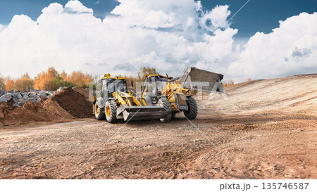 Loaders and excavators are performing earth moving and site preparation at a construction site during the day. The ground is uneven and dusty 135746587
