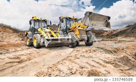 Construction site shows dry, cracked earth with dirt mounds. Two loaders work on the ground while clouds cover part of the sky Construction site shows dry, cracked earth with dirt mounds. Two loaders work on the ground while clouds cover part of the sky 135746589