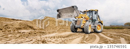 A wheel loader moves across a sandy area, leaving deep tracks. A large pile of sand is visible in the background, indicating ongoing construction work 135746591