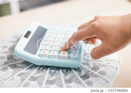 Hand pressing a pastel calculator on scattered US dollar bills, calculating budget, expenses, savings and cash flow for personal finance, investment planning and cost control at home or office. 135746666