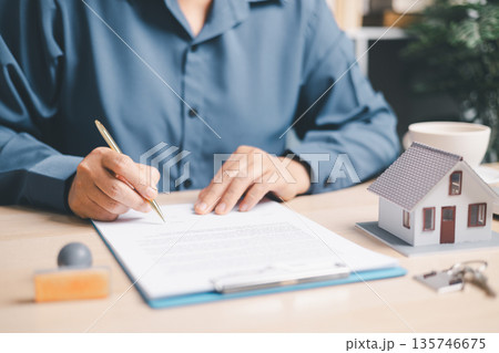 Person signing a real estate contract on clipboard beside a house model and keys, closing a home purchase, mortgage or rental agreement, property insurance and loan approval at office desk. Person signing a real estate contract on clipboard beside a house model and keys, closing a home purchase, mortgage or rental agreement, property insurance and loan approval at office desk. 135746675