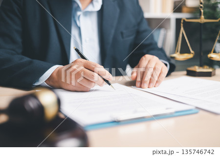 Lawyer in suit signing a contract on clipboard at office desk with justice scales in background, representing legal services, compliance review, agreement approval and documentation. Lawyer in suit signing a contract on clipboard at office desk with justice scales in background, representing legal services, compliance review, agreement approval and documentation. 135746742