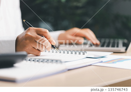Business person taking notes in a spiral notebook beside a laptop and financial charts, planning strategy, budgeting and reporting for corporate analysis, meeting prep and productivity. Business person taking notes in a spiral notebook beside a laptop and financial charts, planning strategy, budgeting and reporting for corporate analysis, meeting prep and productivity. 135746743