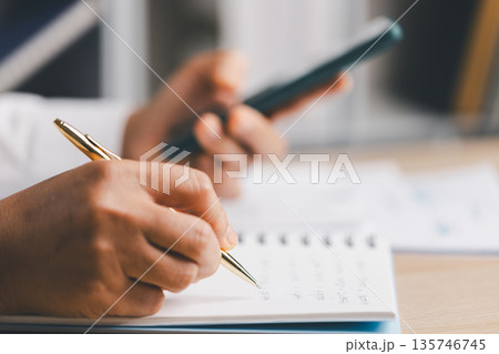Close-up of hands writing notes in a notebook while holding a smartphone, organizing tasks, budgeting and tracking expenses for business productivity, planning and remote work communication. Close-up of hands writing notes in a notebook while holding a smartphone, organizing tasks, budgeting and tracking expenses for business productivity, planning and remote work communication. 135746745