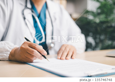 Doctor in white coat with stethoscope signing medical paperwork on clipboard at clinic desk, completing patient records, insurance forms, prescription approval and healthcare documentation. Doctor in white coat with stethoscope signing medical paperwork on clipboard at clinic desk, completing patient records, insurance forms, prescription approval and healthcare documentation. 135746748