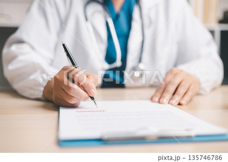 Doctor in white coat with stethoscope signing an approved medical form on clipboard at desk, representing healthcare documentation, prescription, insurance claims and patient care. Doctor in white coat with stethoscope signing an approved medical form on clipboard at desk, representing healthcare documentation, prescription, insurance claims and patient care. 135746786