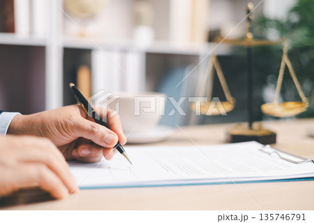 Lawyer in suit signing a contract on clipboard at office desk with justice scales in background, representing legal services, compliance review, agreement approval and documentation. Lawyer in suit signing a contract on clipboard at office desk with justice scales in background, representing legal services, compliance review, agreement approval and documentation. 135746791