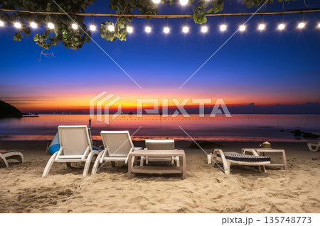 Relaxing beachfront lounge at night with white chairs, decorative string lights, and a vibrant blue and orange twilight sky, Koh Samed Relaxing beachfront lounge at night with white chairs, decorative string lights, and a vibrant blue and orange twilight sky, Koh Samed 135748773