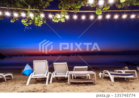 Relaxing beachfront lounge at night with white chairs, decorative string lights, and a vibrant blue and orange twilight sky, Koh Samed Relaxing beachfront lounge at night with white chairs, decorative string lights, and a vibrant blue and orange twilight sky, Koh Samed 135748774