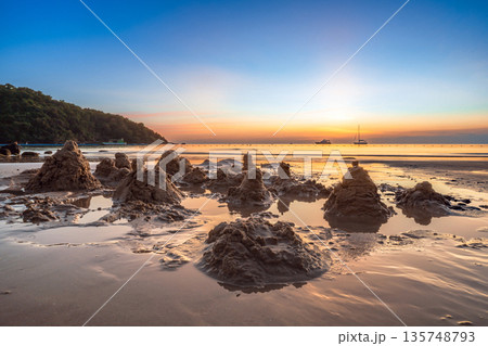 Hand-built sandcastles on a wet beach at twilight with the setting sun reflecting on the water and a serene island atmosphere Hand-built sandcastles on a wet beach at twilight with the setting sun reflecting on the water and a serene island atmosphere 135748793