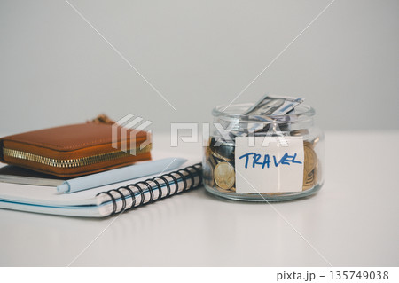 Glass jar labeled TRAVEL packed with coins and banknotes beside notebook, pen and wallet on white background, representing vacation savings, trip budgeting and expense tracking goals. Glass jar labeled TRAVEL packed with coins and banknotes beside notebook, pen and wallet on white background, representing vacation savings, trip budgeting and expense tracking goals. 135749038